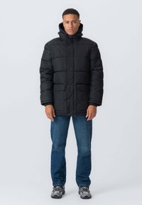 Sort pufferjakke med hætte, quiltet design, fors клапlåse og to frontlommer, parret med blås jeans og sneakers.
