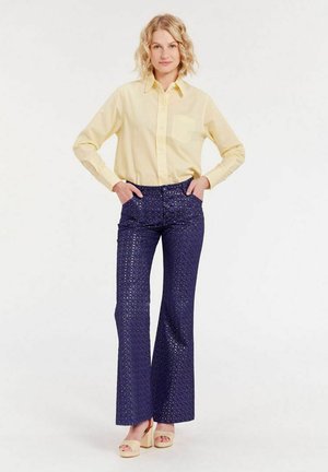 Chemise jaune à manches longues avec une poche poitrine, associée à un pantalon bleu marine évasé avec un motif texturé, et des sandales à plateforme de couleur claire.