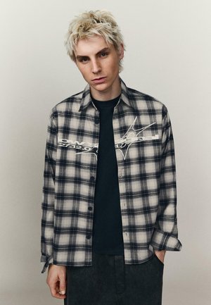 Młody mężczyzna z wybielonymi włosami nosi czarno-białą koszulę w kratę na czarnej koszulce, stojąc na tle prostego jasnego tła.