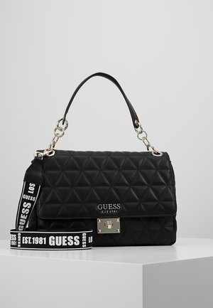 Sac à main matelassé noir Guess avec poignée supérieure, fermoir argenté et bandoulière de marque amovible reposant sur une surface blanche.
