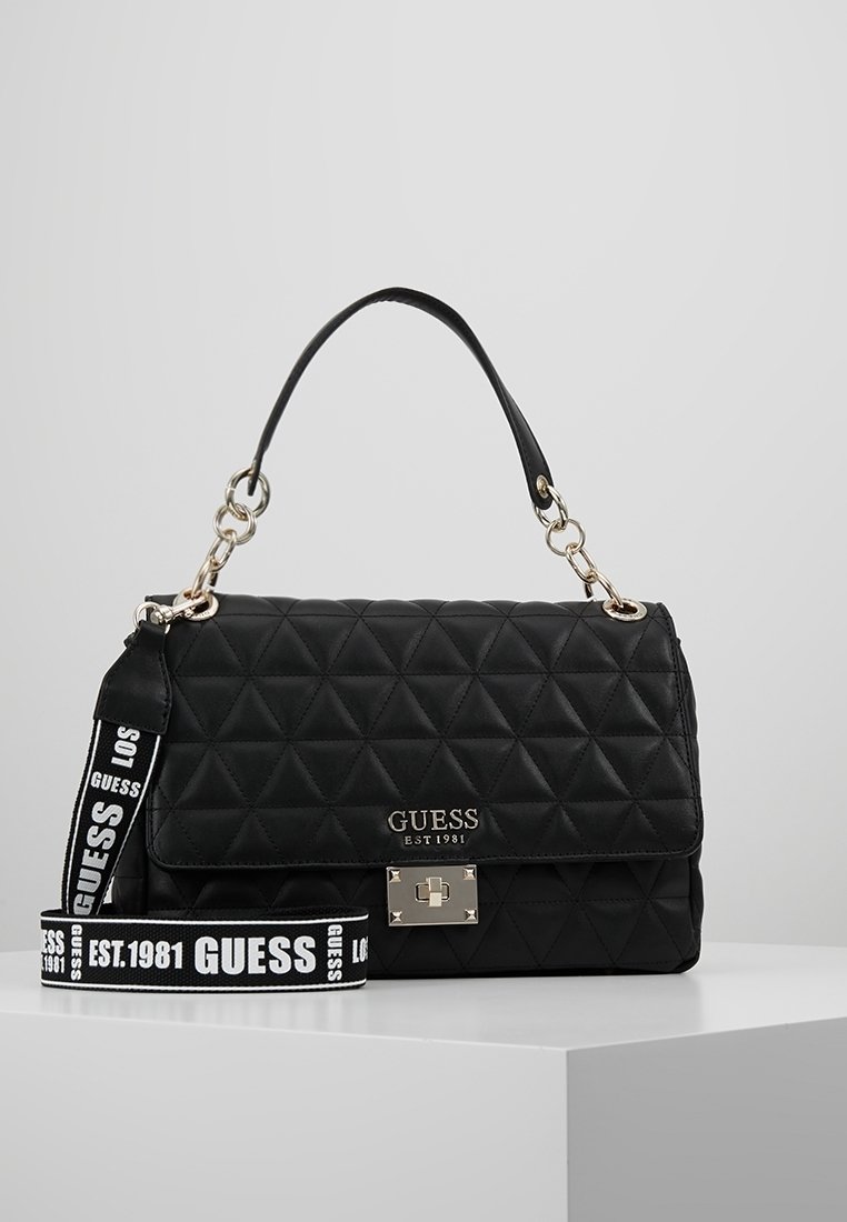 Sac à main matelassé noir Guess avec poignée supérieure, fermoir argenté et bandoulière de marque amovible reposant sur une surface blanche.