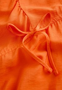 Orange tyg med en slät textur, som har en knuten rosett och veckad sömnad i tyget.