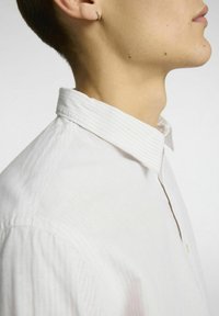 Balta juostuota marškiniai su sulankstytu apykakle, lengva medžiaga, matoma tekstūra ir viena sagė ant apykaklės, dėvimi asmens.