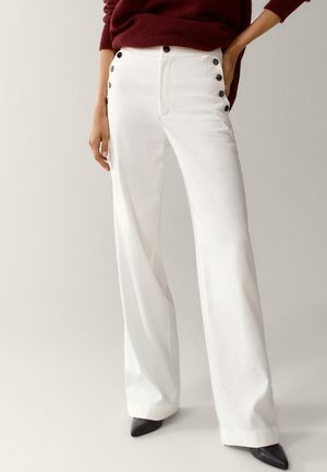 Witte broek met brede pijpen van gladde stof, voorzien van zwarte knoopaccenten langs de zijkanten, gecombineerd met zwarte, puntige hakken.