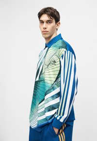 Junger Mann trägt ein langärmeliges Poloshirt von Adidas in Blau, Grün und Weiß mit abstrakten und gestreiften Mustern, Hände in den Taschen einer blauen Hose.