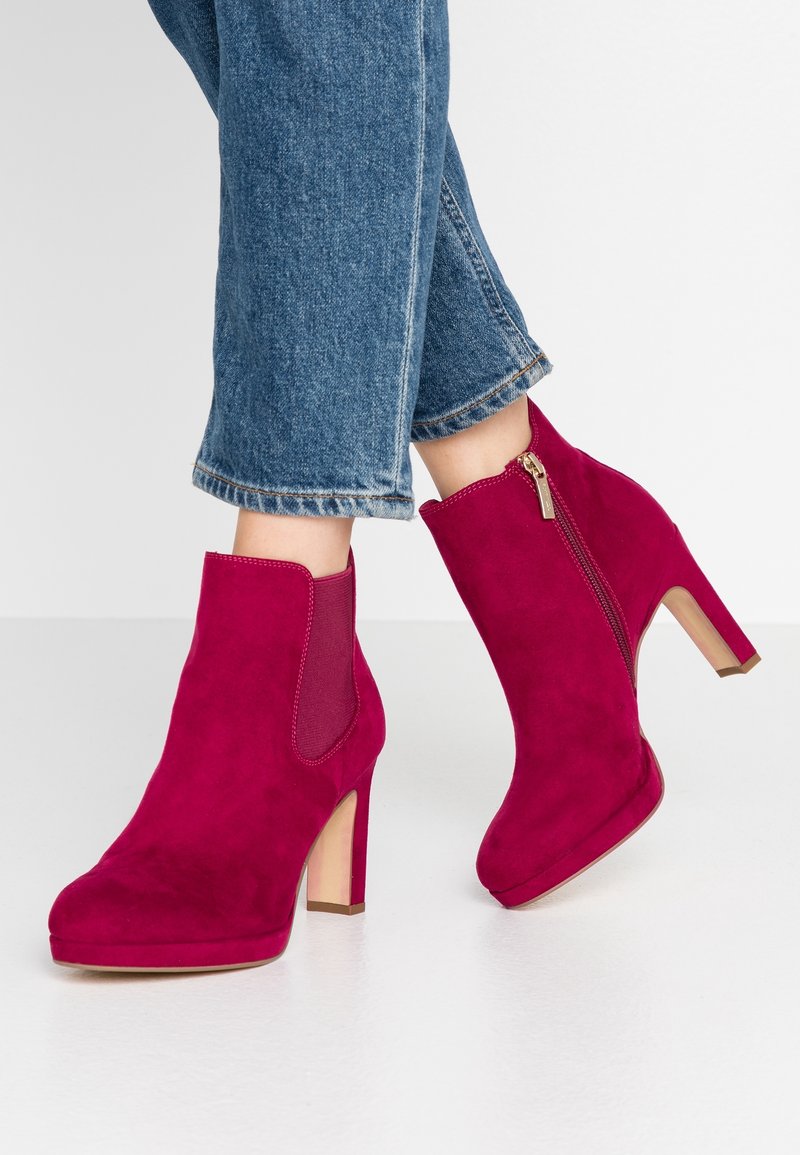 Femme portant un jean bleu et des bottines à talons hauts en daim magenta avec fermeture éclair latérale et panneau élastique, debout sur une surface blanche.
