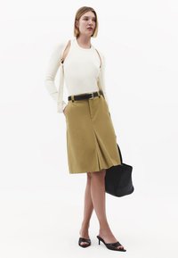 Mujer con top sin mangas beige y cárdigan sobre los hombros, falda kaki hasta la rodilla, cinturón negro, lleva bolso negro y zapatos de tacón negros.