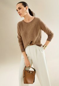 Eine Frau trägt einen braunen strukturierten Pullover und weiße Hosen, während sie eine braune Lederhandtasche hält und mit goldenen Ohrringen und einem Armband accessoriert ist.