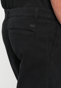 Black cotton trousers featuring a textured finish, side and back pockets, and a small black fabric label with "BOSS" stitched on.