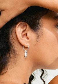 Boucle d'oreille en argent présentant un design de main sculptural et un détail de pointe, suspendue à un anneau classique. Texture métallique lisse.