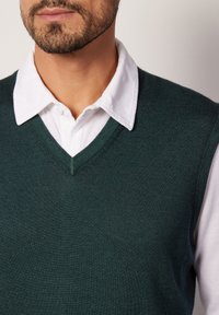 Ciemnozielony sweter z dzianiny z dekoltem w kształcie litery V, noszony na białej koszuli z kołnierzykiem. Gładka tekstura, precyzyjne szwy oraz warstwowy wygląd.