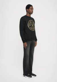 Črna majica s kapuco z zlatim okroglim logotipom spredaj, kombinirana s temno sivo džinsom in črnimi športnimi copati. Gladek material, sproščen kroj.