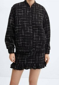 Personne portant une veste en tweed noire et une jupe mini assortie avec un motif en quadrillage blanc et un ourlet froncé, se tenant devant un fond uni.