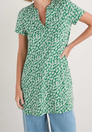 Kobieta w zielonej tunice z krótkim rękawem z białym nadrukiem stokrotek i dekoltem w serek, w połączeniu z jasnoniebieskimi dżinsami, jedną ręką na biodrze, na jednolitym tle.
