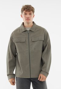 Zielona oliwkowa kurtka zapinana na zamek błyskawiczny z kołnierzem, dwoma kieszeniami na piersi i detalami na guzikach. Wykonana z tkaniny o gładkiej fakturze.
