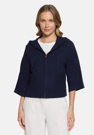 Woman wearing a navy blue zip-up hoodie with three-quarter sleeves paired with white pants, standing against a light background.