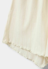 Világos krémszínű szövet felső, függőleges csíkokkal, diszkrét textúrával és hullámvonalú alsó szegéllyel, apró fodrokkal díszítve.