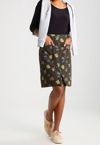 Falda de color verde oscuro con un estampado floral y bolsillos. Hecha de tejido texturizado, de longitud hasta la rodilla y con corte recto. Combinada con una blusa negra ajustada y zapatillas beige.