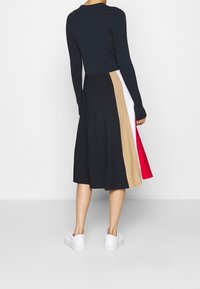 Zwart lange mouw jurk met een geplooide, multicolore rok in beige, wit, rood en zwart. Witte sneakers maken de look compleet.