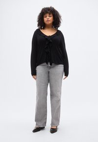 Blouse noire à volants sur le devant avec manches longues, associée à un jean gris clair à jambes droites et des ballerines noires pointues. Textures lisses sur l'ensemble.