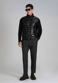 Puffer vest van glanzend zwart materiaal, langwerpige zwarte trui eronder, grijze op maat gemaakte broek en donkerbruine schoenen. Stijlvolle zonnebril inbegrepen.