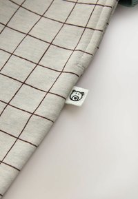 Material gri cu un model de grilaj maro, având un mic etichetă cu logo-ul unui urs pe margine. Textură ușoară și netedă, îndoită la colț.