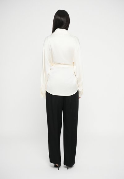 Camicia di seta color crema con colletto e maniche lunghe, abbinata a pantaloni neri a vita alta con taglio dritto.