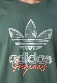 Zelené bavlněné tričko s velkým stříbrným logem Adidas a třemi pruhy, na kterém je oranžový text "Originals". Kulatý výstřih, krátké rukávy.