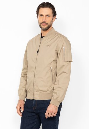 Mann mit Bart, der eine beigefarbene Bomberjacke mit Reißverschlusstaschen und dunkelblaue Jeans trägt, steht vor einem schlichten weißen Hintergrund.