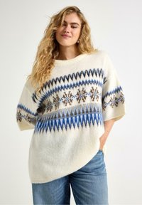 Maglione crema con motivi geometrici blu e marroni, vestibilità rilassata, maniche corte e finitura morbida e testurizzata. Abbinato a jeans in denim blu.