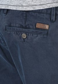 Pantalons bleu marine en tissu lisse, dotés d'une poche avant avec un label en cuir marron et une fermeture à bouton.