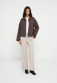 Καφέ μάλλινο μπουφάν oversized με γιακά, τσέπες με καπάκι και κλείσιμο με φερμουάρ, σε συνδυασμό με ανοιχτό μπεζ παντελόνι και μαύρα μποτάκια με τακούνι.