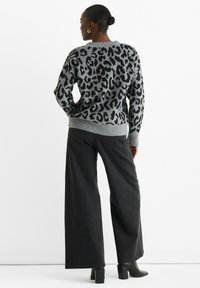 Grauer Pullover mit schwarzem Leopardenmuster, Rundhalsausschnitt und gerippten Bündchen. Kombiniert mit schwarzen Schlagjeans und schwarzen Stiefeln mit Absatz.