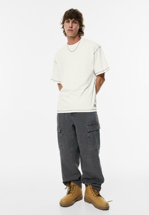 Weißes T-Shirt mit kurzen Ärmeln und kontrastierenden Nähten, graue Cargo-Hose mit mehreren Taschen und sandfarbene Schnürstiefel. Schlichtes, sauberes Design.