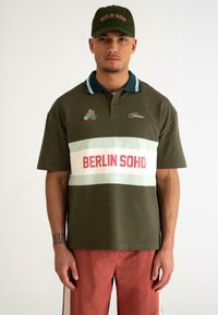 Oliven grøn polo skjorte med kontrasterende lyseblå og hvide vandrette striber; "BERLIN SOHO" i fed rød over brystet; broderede detaljer.