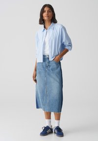 Camicia button-up a righe blu chiaro sopra una maglietta bianca, abbinata a una gonna di denim e sneaker blu. La gonna presenta una cucitura frontale e un orlo sfrangiato.