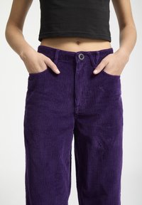 Pantalons en velours côtelé violet avec des côtes verticales, dotés de poches latérales et d'une fermeture à boutons à la taille. Le tissu a une texture douce.