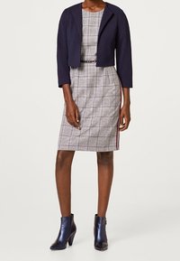 Giacca corta blu navy sopra un vestito a quadri con cintura, caratterizzata da una cerniera laterale rossa. Abbinata a stivaletti con tacco blu scuro.