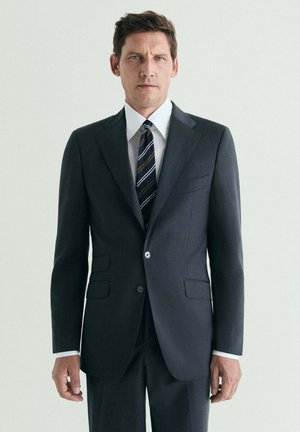 Hombre que lleva un traje gris oscuro, camisa blanca y corbata de rayas, de pie frente a un fondo claro y liso.