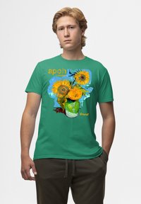 Camiseta de algodón verde con un estampado gráfico de girasoles en un jarrón y el texto "apoh London Vincent" en naranja y azul.