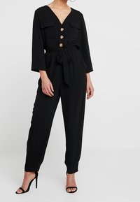 Svart jumpsuit med V-ringning, två framfickor, gulddetaljer, midjeband och avsmalnande ben. Bärs med svarta höga sandaler.