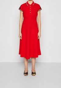 Rode polojurk van katoen met een uitlopende rok. Voorzien van een knoopsluiting, korte mouwen met contrasterende afwerking en gedragen met zwarte schoenen.