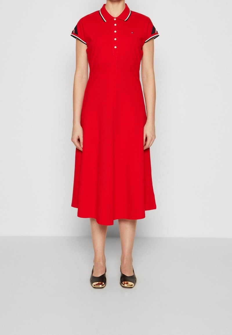 Rode polojurk van katoen met een uitlopende rok. Voorzien van een knoopsluiting, korte mouwen met contrasterende afwerking en gedragen met zwarte schoenen.