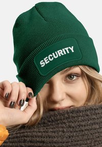 Green knit beanie with a stitched patch displaying "SECURITY" in bold white letters; textured ribbed pattern; close-up on the model's face.
