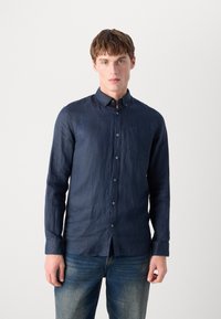 Navy blue, long-sleeve shirt made of linen, featuring a button-down collar, front pocket, and subtle texture. Paired with blue jeans.