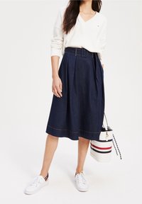 Jupe midi en denim, bleu foncé, design plissé, taille ceinturée, détails de couture blanche à l'ourlet. Associée à des baskets blanches et un sac rayé.