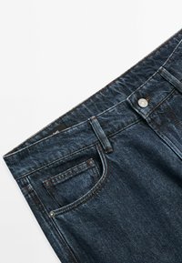 Mörka denimjeans med en slät yta. Funktioner inkluderar en framficka, kontrasterande sömmar och en metallknäppning.