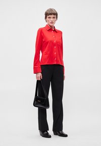 Κόκκινο μακρυμάνικο πουκάμισο με κουμπιά και γιακά, συνδυασμένο με μαύρα φαρδιά παντελόνια και μαύρες μπότες με τακούνι; κρατώντας μια μικρή μαύρη τσάντα.