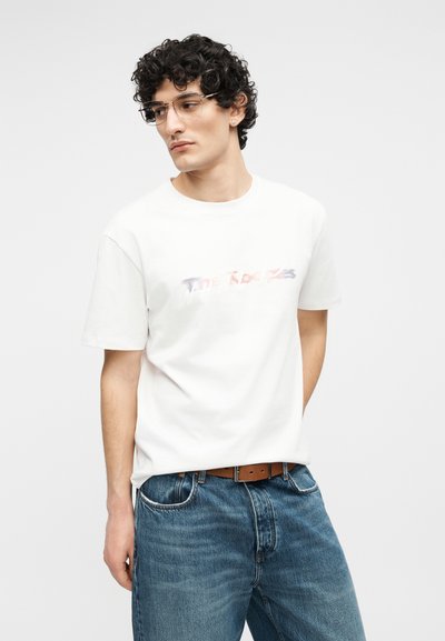 Νεαρός με σγουρά μαλλιά και γυαλιά φοράει λευκό γραφικό μπλουζάκι και μπλε τζιν με καφέ ζώνη, κοιτώντας προς τα αριστερά.