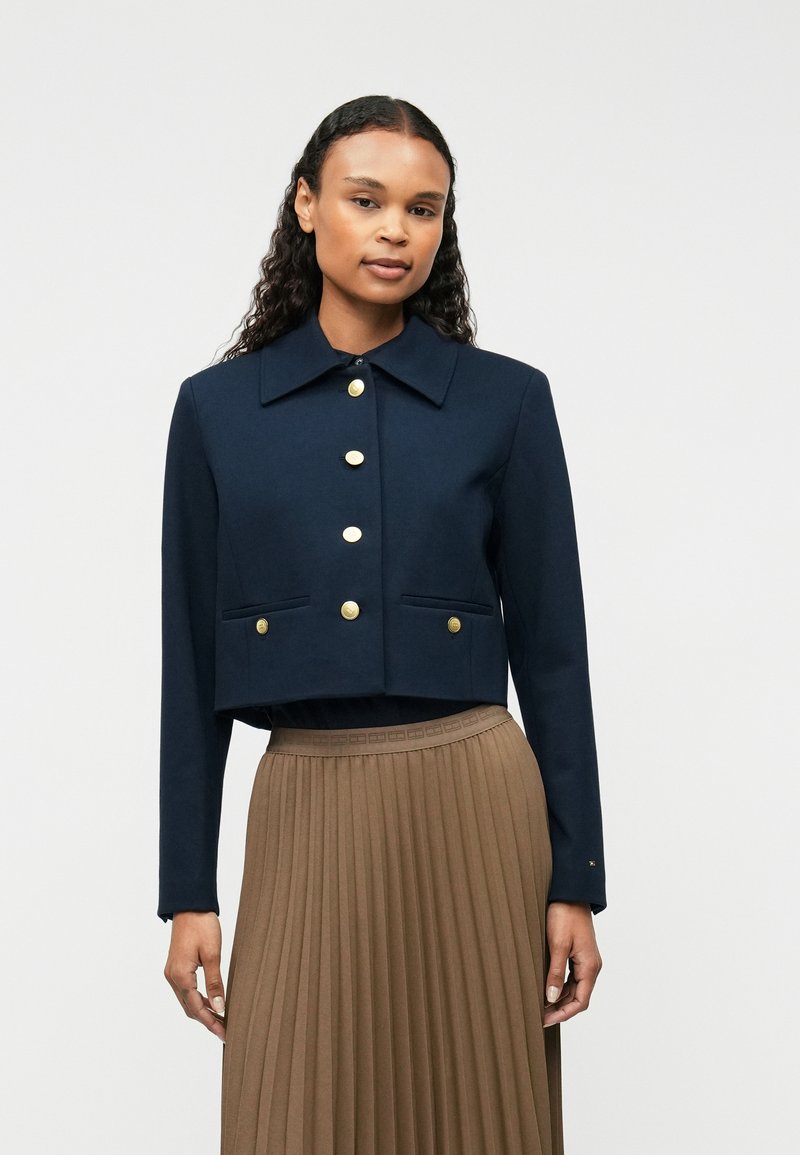 Kobieta ubrana w granatową krótką kurtkę z złotymi guzikami oraz brązową, plisowaną spódnicę z wysokim stanem, stojąca na jednolitym tle.